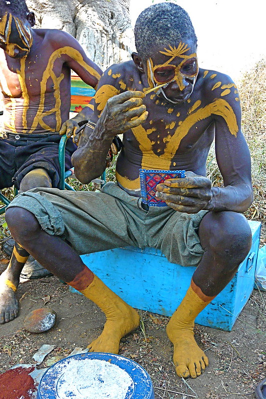 A man sits on a blue box, painting designs on his face with yellow and black paint.